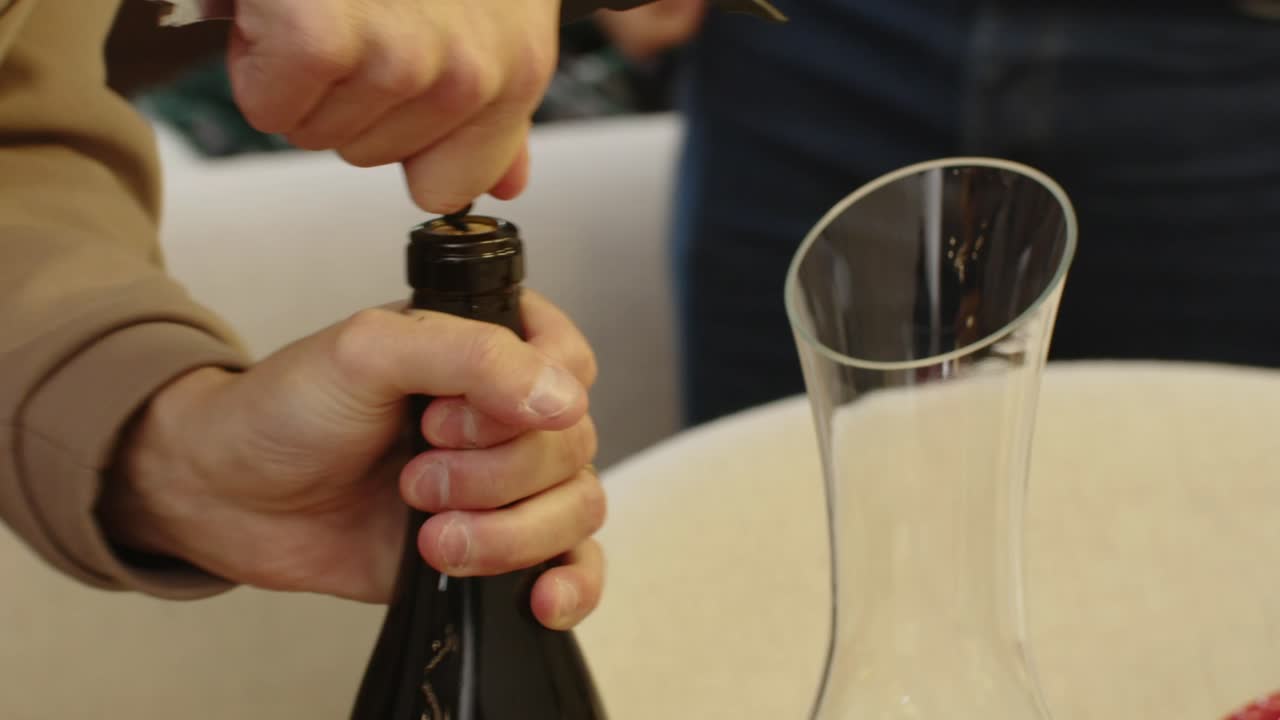 Hands opening a wine bottle with a corkscrew beside a glass decanter