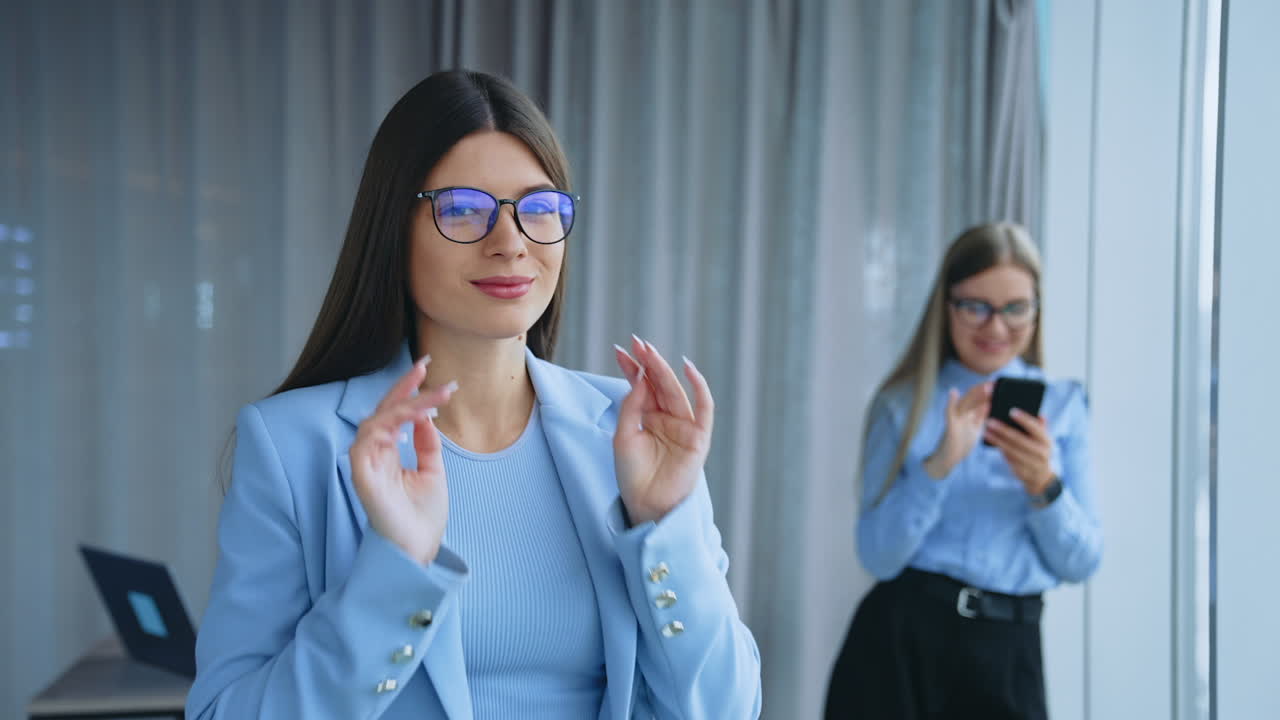 Good-looking young long-haired lady wearing blue jacket stands at the desk in office. Business woman puts on glasses smiling to camera. Another lady holding phone standing at window at backdrop in blur.