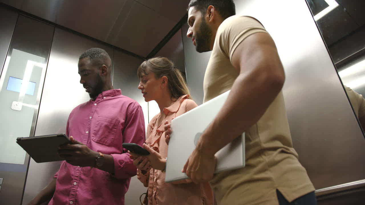 Collaborating in elevator, diverse colleagues using tablet, smartphone, and laptop for work