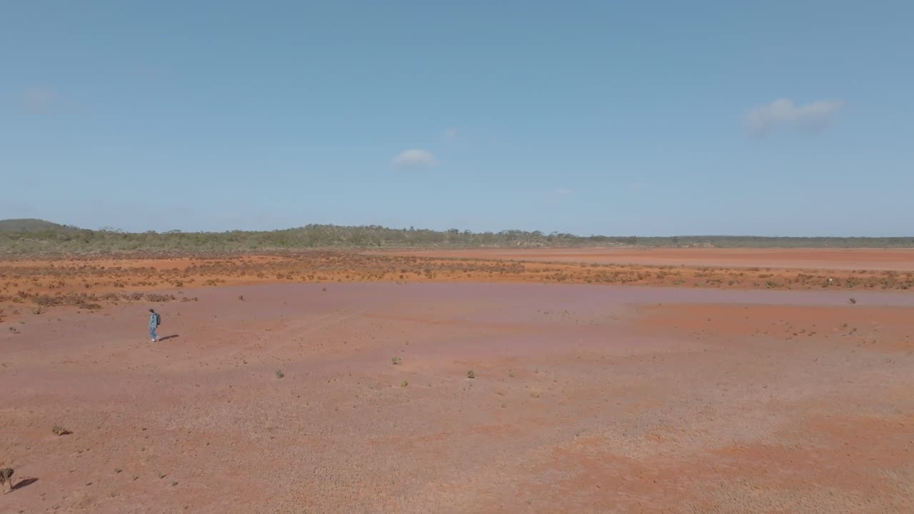 Panning drone clip from right to left, showing rugged Australian outback habitat and vegetation, and male backpacker walking on baked orange desert earth