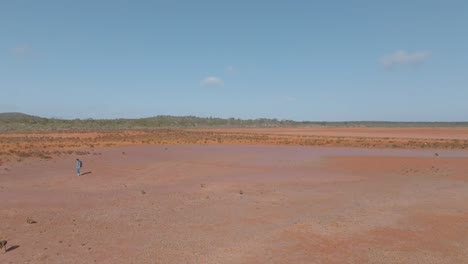 Panning-drone-clip-from-right-to-left,-showing-rugged-Australian-outback-habitat-and-vegetation,-and-male-backpacker-walking-on-baked-orange-desert-earth