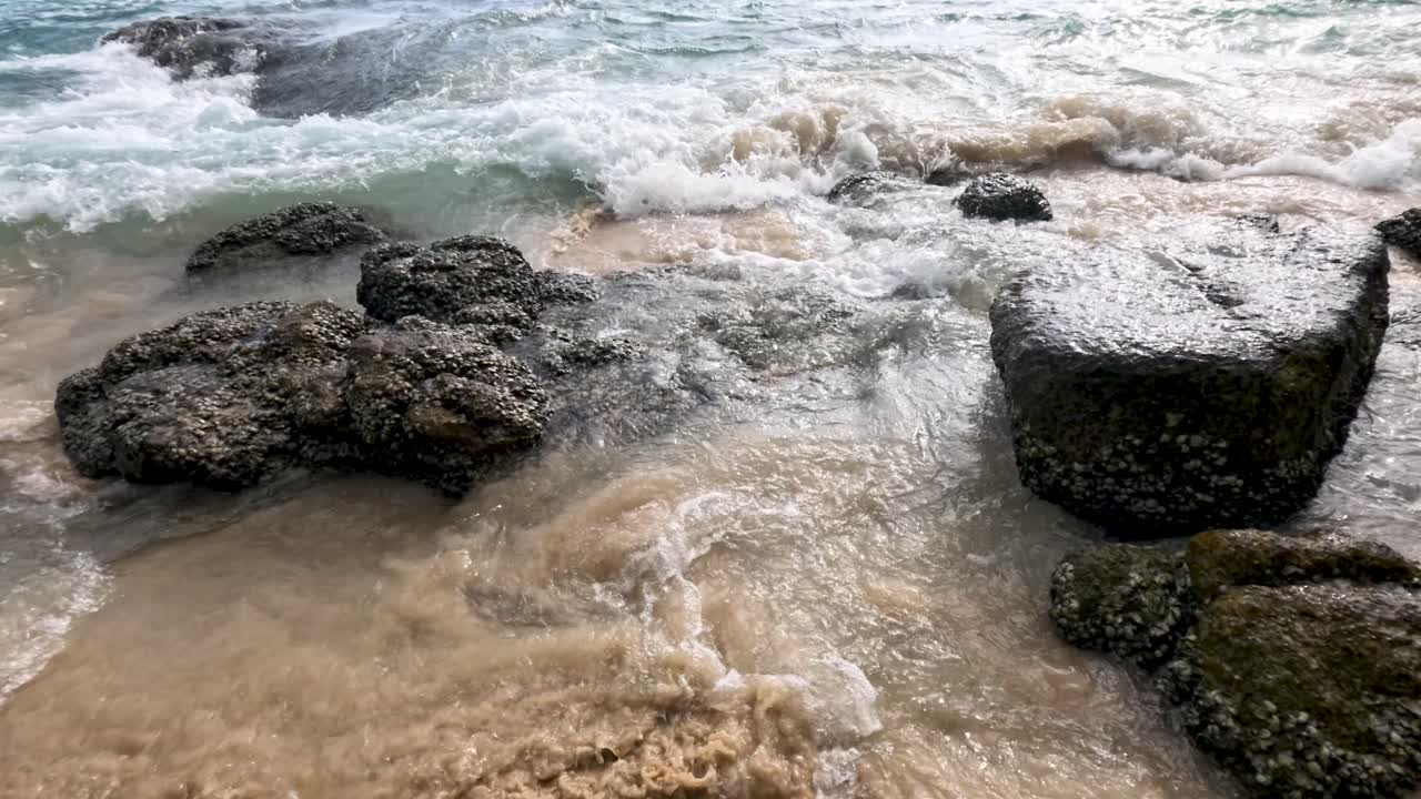 Ocean waves surge over textured rocks and sand, daylight, handheld camera, dynamic natural movement