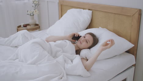 young caucasian woman sitting on the bed in the morning feels bad. a young woman is making a smartphone call to her doctor.