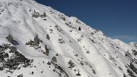 Drone-shot-tracking-from-left-to-right-along-a-snow-covered-mountain-side,-with-interesting-snow-formations-and-rocks