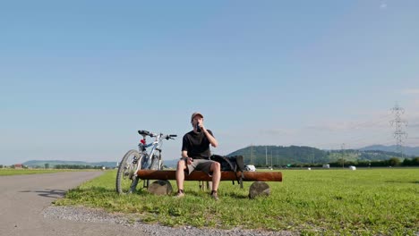 Junger-Männlicher-Radfahrer-Macht-An-Einem-Sonnigen-Tag-Mit-Klarem-Blauen-Himmel-Im-Hintergrund-Eine-Pause-Auf-Einer-Parkbank-Und-Trinkt-Wasser-Aus-Der-Flasche