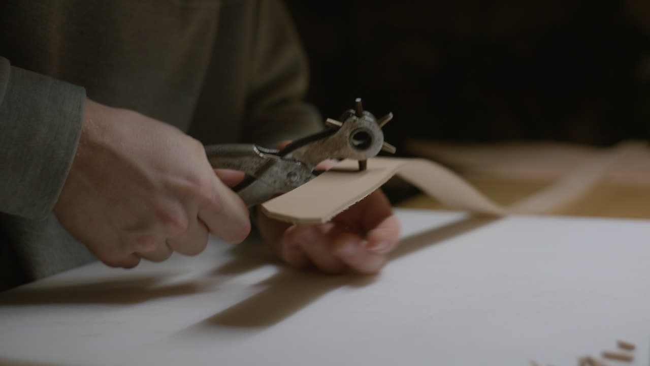 A craftsman presses a rotary hole punch onto a leather strap, carefully preparing it for stitching or fastening.