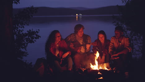 friends playing guitar around campfire by lake at night