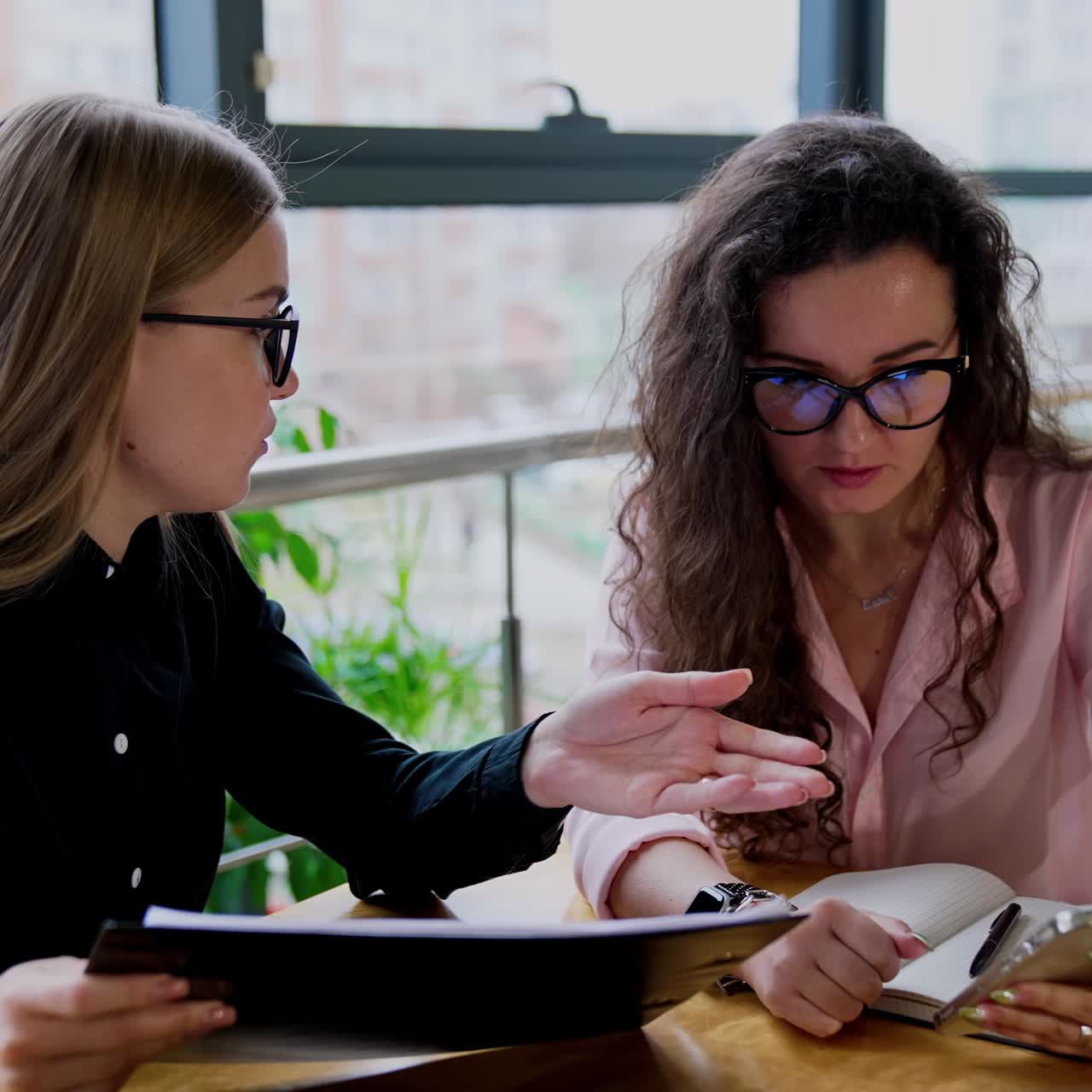 Mid-aged young business ladies discussing job issues. Long-haired women in glasses sitting at desk looking at phone and communicating