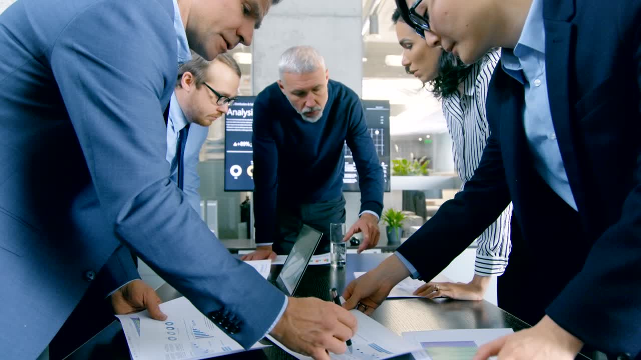 nella sala riunioni un gruppo diversificato di uomini d'affari in piedi attorno al tavolo che hanno una sessione di pianificazione strategica. lavorano su laptop, scambiano appunti e idee.