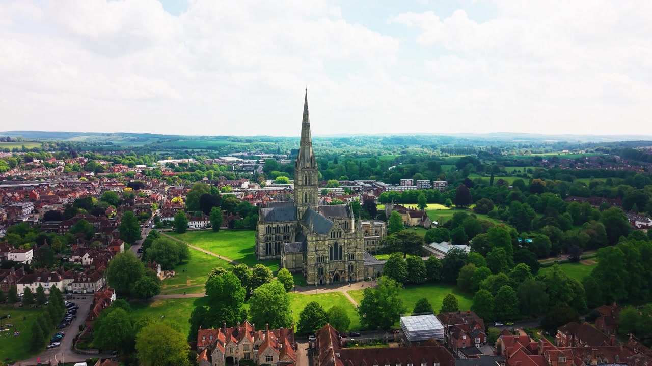 Riprese aeree con drone della Cattedrale di Salisbury nel Wiltshire, che mostrano la famosa guglia della cattedrale, il centro storico, il Cathedral Close, il parco verde, i tetti, gli alberi e la campagna circostante.
