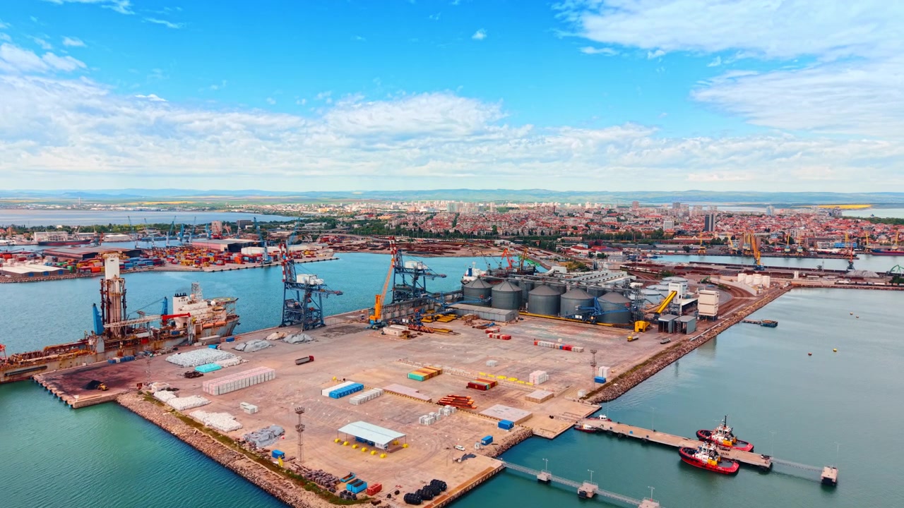 Aerial view of cargo dock with maritime vessel and tugboats. Industrial port terminal featuring specialized maritime vessel, tugboats, and grain silos on concrete pier.