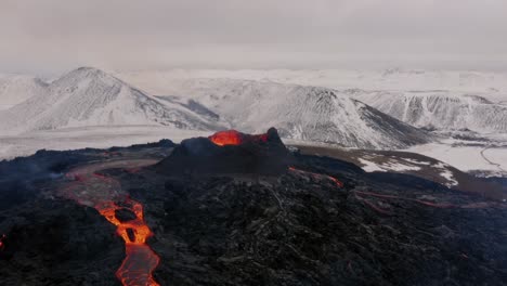 Mit-Einer-4K-Drohne-Aufgenommene-Luftaufnahmen-Zeigen-Einen-Ausbrechenden-Vulkan,-Der-Lava-Aus-Seinem-Krater-Spuckt,-Mit-Dramatischen-Berggipfeln-Der-Alpen-Im-Hintergrund