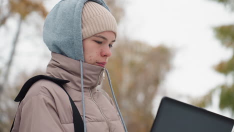 young light-skinned woman wearing layered hoodie and jacket looks attentively at laptop screen while standing outdoors on snowy day, with blurred autumn trees and serious facial expression