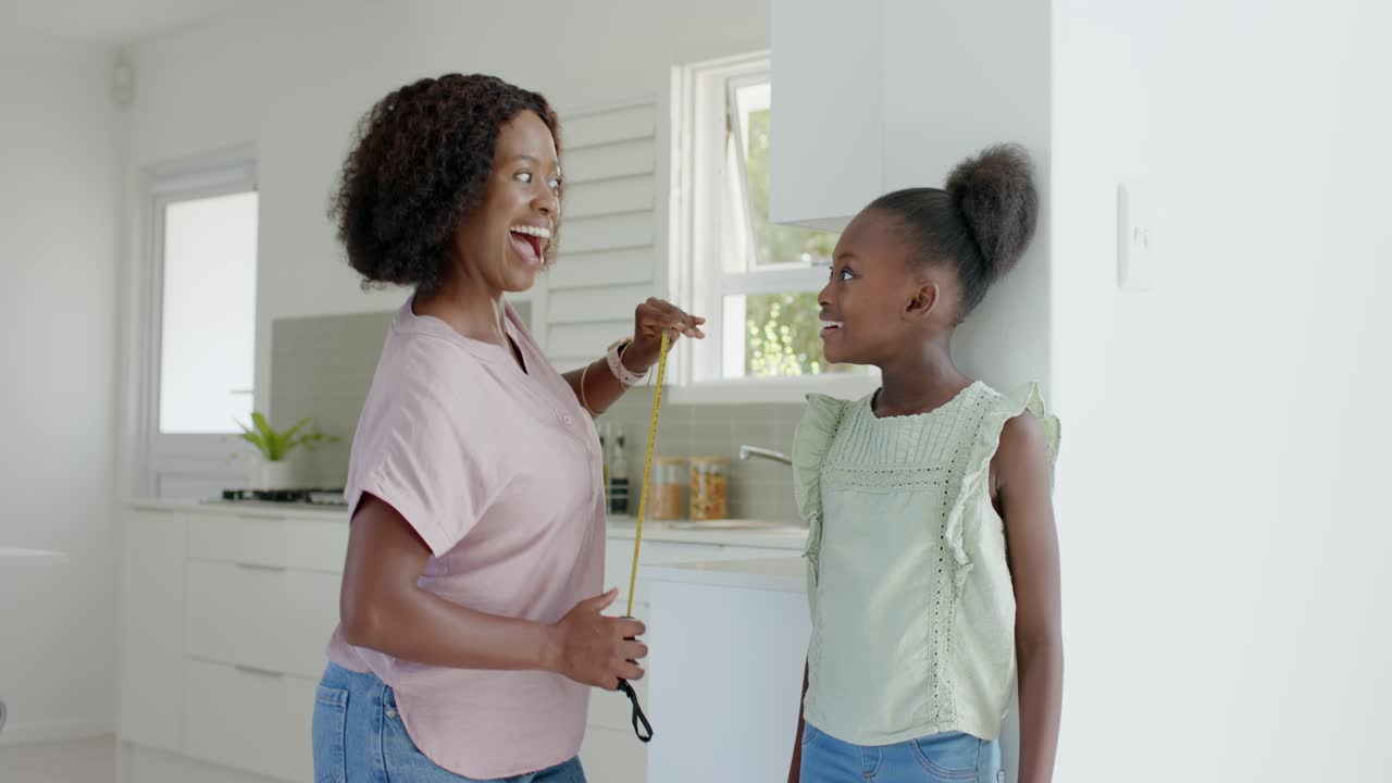African American mom placing yellow tape at daughter's head, measuring height against kitchen wall