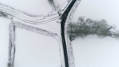 Draufsicht-Auf-Eine-Schneebedeckte-Landschaft-Mit-Einer-Geräumten-Asphaltstraße-Und-Einer-Abzweigung,-Die-Mit-Schnee-Bedeckt-Und-Mit-Sichtbaren-Autoreifenspuren-Bedeckt-Ist