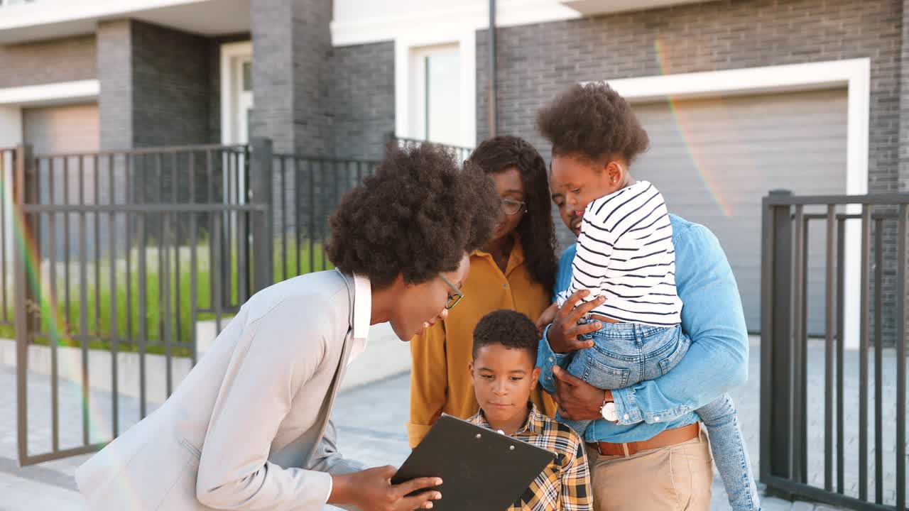 Cheerful African American family with small kids buying house at suburbs and talking with female real-estate agent outside