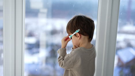 rear view of a boy standing at the window wearing sunglasses. cropped image of a toddler looking into the window on sunny daytime.
