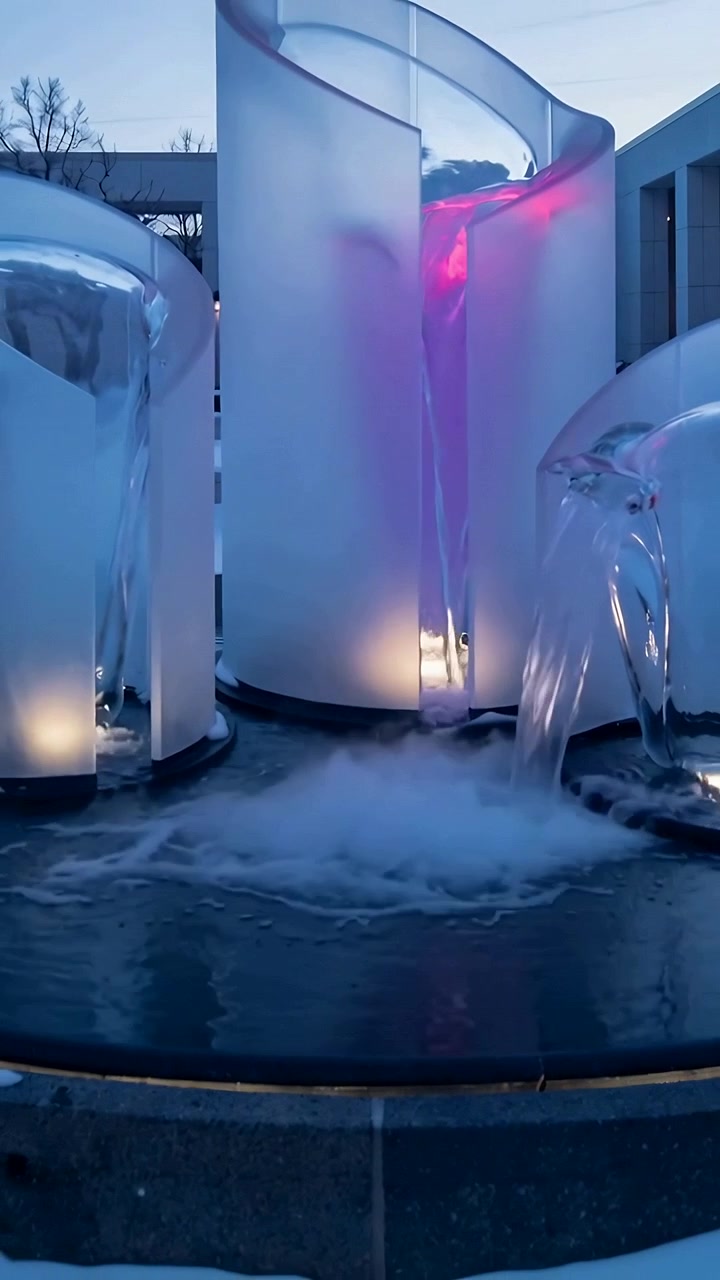 Vertical video: Pumps engaging fountain pouring from spouts into basin at dusk lit panels misting