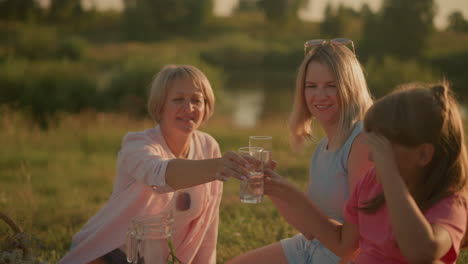 family enjoying outdoor picnic, smiling as elderly woman hands glass of water to daughter, spending quality time together under warm sunlight, with another woman holding water close by