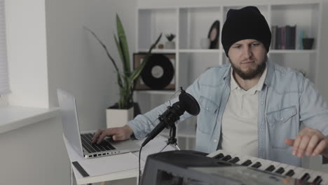 Male-Musician-Playing-Electric-Keyboard-And-Using-A-Laptop-At-Home