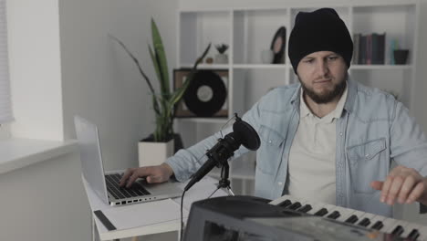 músico masculino tocando teclado eléctrico y usando una laptop en casa