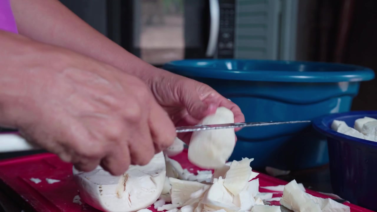 Mani femminili che usano un coltello per sbucciare e tagliare radici di manioca fresca su un tagliere rosso. Preparazione di cibo rurale per la conservazione domestica