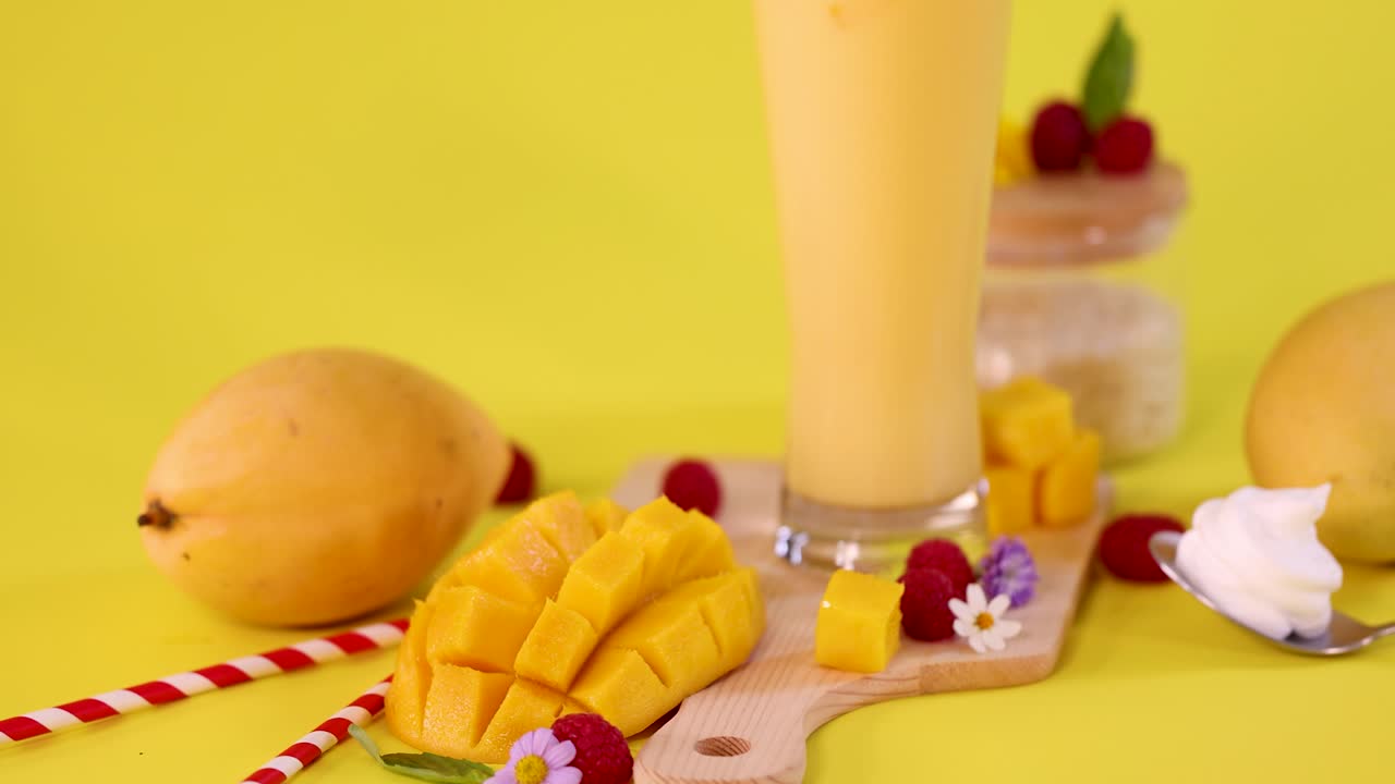Tall mango smoothie, fresh mango cubes, raspberries, whipped cream, bright yellow background, static camera