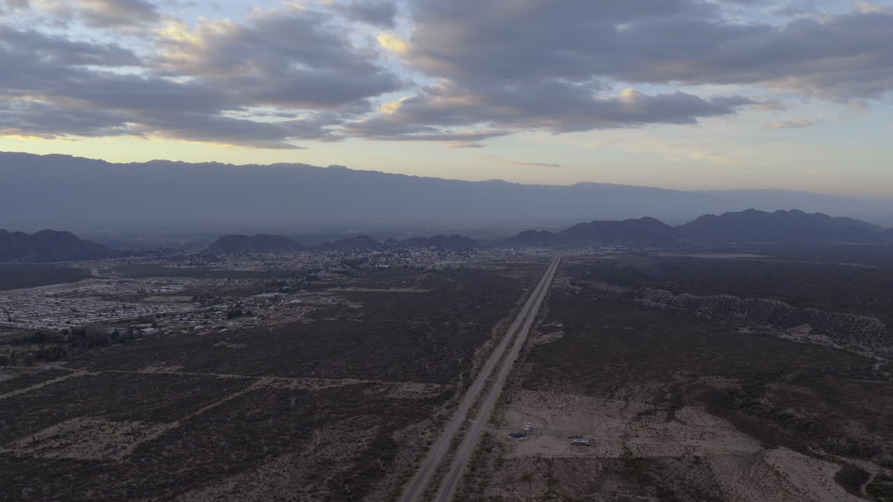 Un drone vola in avanti sopra una lunga strada dritta all'alba, con la città e le montagne che sfumano in un orizzonte blu nebbioso