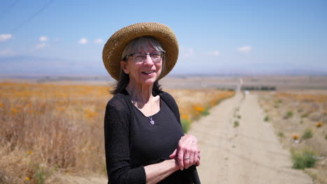 A-happy-middle-aged-woman-with-gray-hair-smiling-on-a-dirt-road-with-orange-poppy-flowers-under-blue-skies-SLOW-MOTION