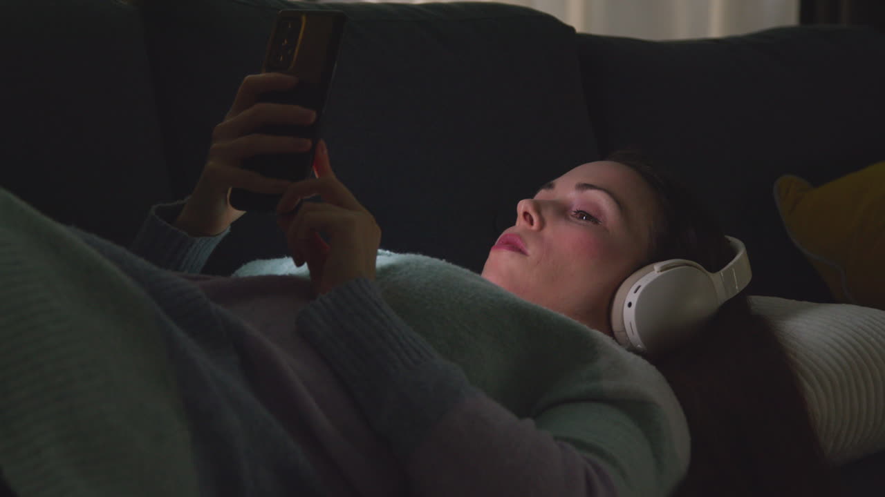 Woman Wearing Wireless Headphones Lying On Sofa At Home At Night Streaming Music Or Watching Movie On Mobile Phone 1