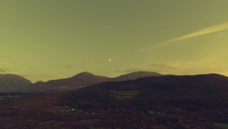 aerial drone flight over forests in north wales towards a half moon rising in the evening sky above snowdonia mountains. scenic landscape view with wilderness, twilight, and natural beauty