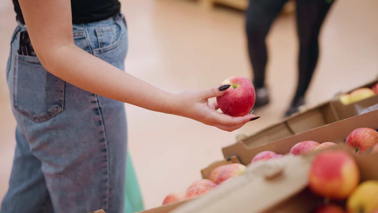 은 사과를 들고 식료품 가게에서 쇼핑하는 여성의 부분적인 시야, 그것을 주의 깊게 관찰하는, 그녀의 주위에 상자에 사과를 가지고 배경에 접근하는 누군가의 흐릿한 시야