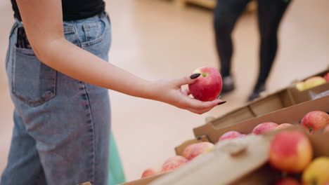 partial view of woman shopping in grocery store holding red apple, observing it carefully, blurred view of someone approaching in background with apples in boxes around her