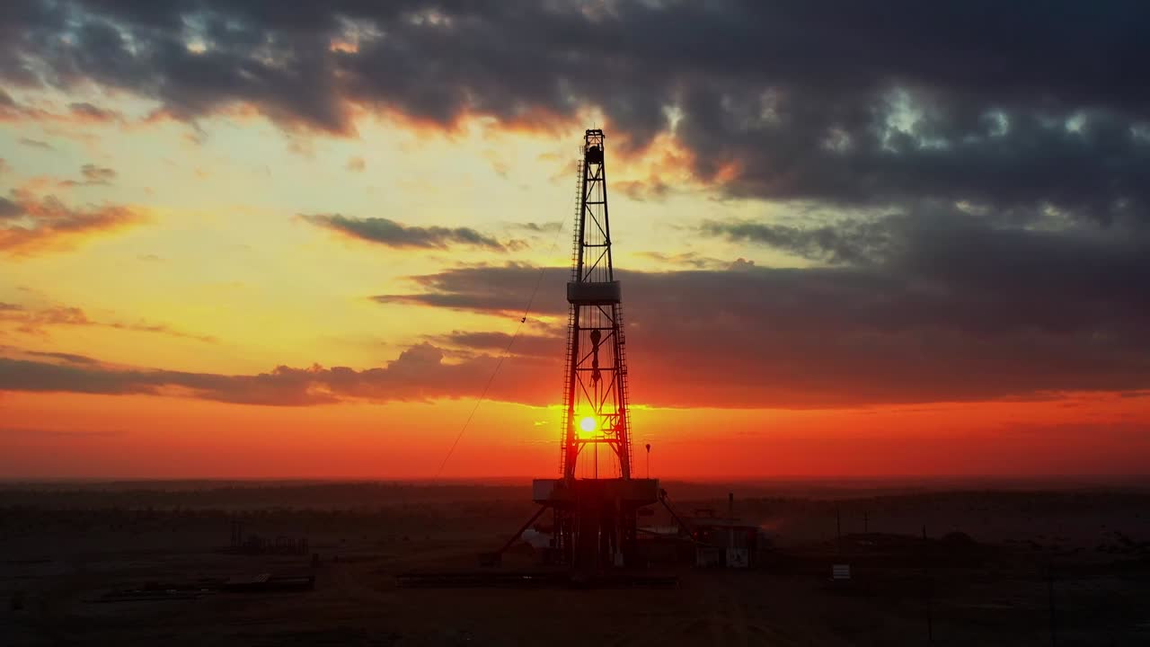Silhuet af olieboreplatform ved solnedgang i ørkenlandskab med dramatisk himmel.