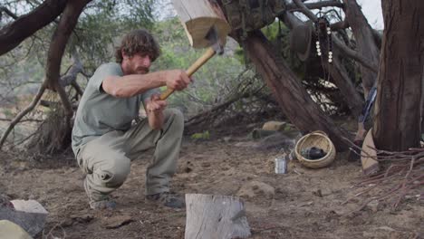 bearded caucasian male survivalist using axe to cut fire logs at camp in wilderness