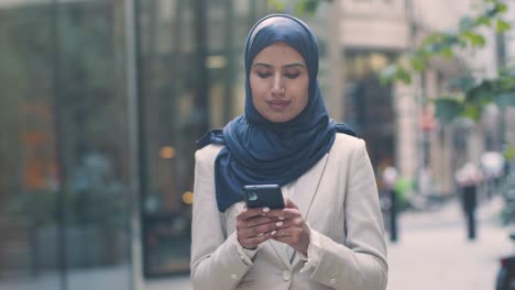 Mujer-De-Negocios-Musulmana-Revisando-Mensajes-En-Un-Teléfono-Móvil-Parada-Afuera-De-La-Oficina-En-La-Ciudad-1