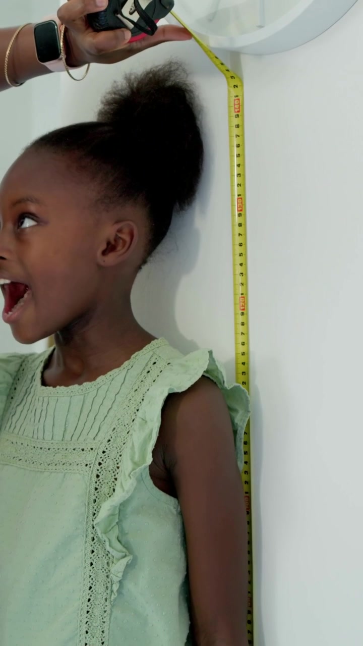 Vertical video: Moving mom holding tape measuring girl in green dress home checking height hugging