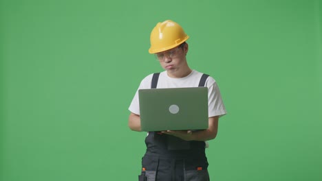 asian man worker wearing goggles and safety helmet using a laptop and having a headache while standing in the green screen background studio