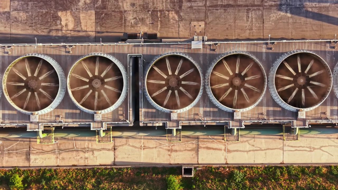 Aerial view shows large cooling towers at an industrial site in sunlight with patterns on the ground and green grass nearby in a clear blue sky