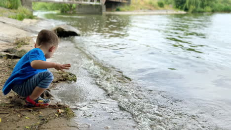 caucasian kid sits squatted on the shore close to the water. baby boy picks and throws back wet sand.