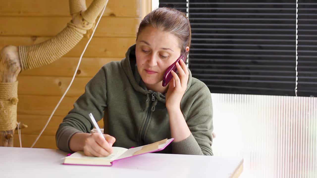 Woman speaking on phone while looking down writing in notebook, tilt down