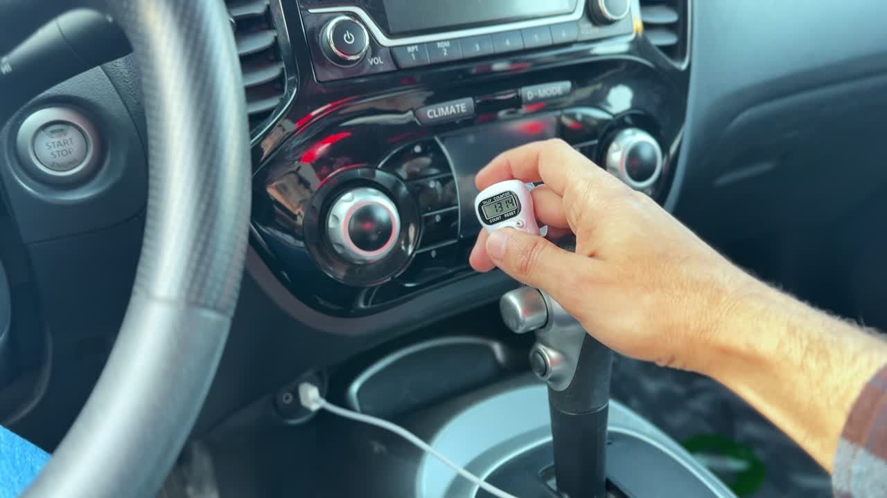 Nahaufnahme einer Fahrerhand, die einen kleinen digitalen Zähler im Innenraum eines modernen Autos in der Nähe des Schalthebels und des Klimabedienfelds hält, was das Konzept von Transport und Zählung verdeutlicht.