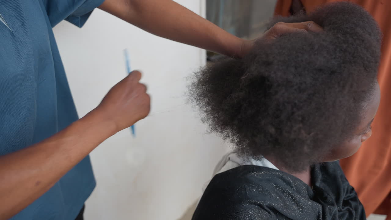 Primo piano di pettine per capelli che mostra lo stilista che districa la texture afro naturale, cliente seduta con mantella protettiva, mani che lavorano delicatamente tra le spirali, luce interna calma, cura attenta del cuoio capelluto, tecnica lenta e precisa.