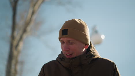 a close-up of an adult wearing a brown jacket and a beige beanie, talking while walking outdoors on a bright, sunny day, with a blurred view of a bare tree and a clear sky