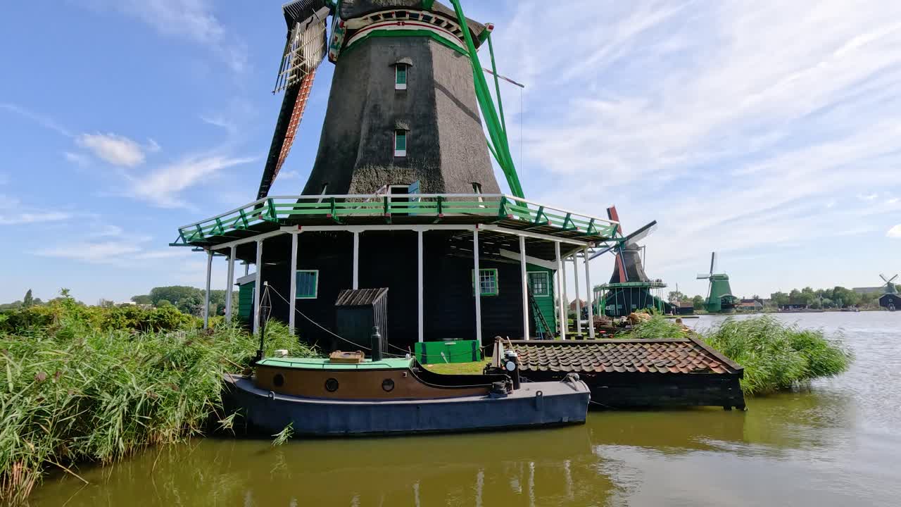 Historic windmill slowly turning by riverside, bright daylight, wide shot, calm summer atmosphere
