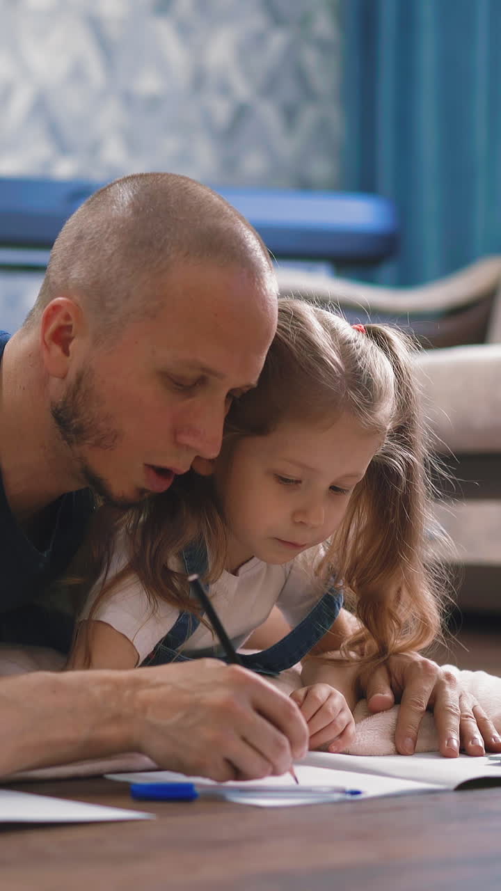 cu, cam tracking: young father and daughter lie on floor in living room, holds small hand, ballpoint pen helping draw in notebook homework
