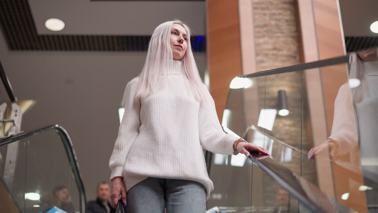 chic woman wearing white sweater and jeans holding handbag descending moving walkway in bright modern mall with blurred family members following behind reflective glass railing overhead lights