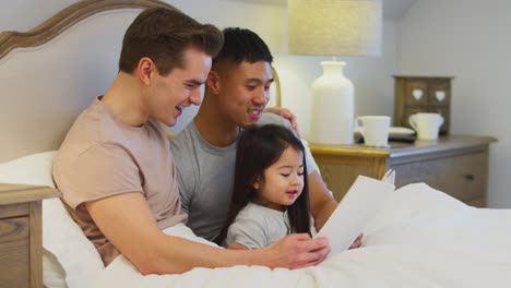 family with two fathers in bed at home reading story to daughter