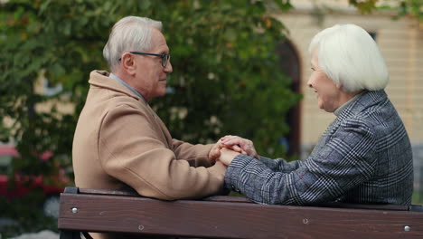 side view of elderly married couple holding hand and talking sitting on the bench in the park in autumn 1