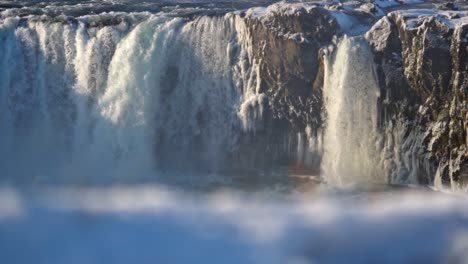 iceland's iconic goðafoss, the "waterfall of the gods," roars powerfully through a serene winter landscape, its cascading waters framed by snow-covered cliffs and icy formations